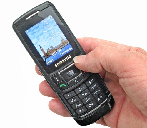 A hand holding a Samsung SGH-D900 black slider mobile phone with screen displaying wallpaper of a cityscape.