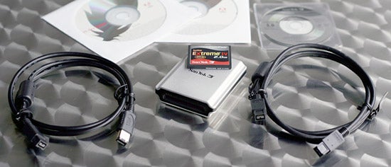 SanDisk Extreme IV 2GB CompactFlash memory card with attached label next to a card reader and two USB cables on a textured grey background, with software CDs partially visible in the background.