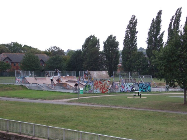 A photograph taken with a Fujifilm Finepix F30 camera featuring a skate park with graffiti-covered ramps and a few skaters in the distance, surrounded by green trees and a walking path.