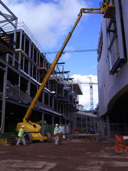 Construction site with a yellow telescopic boom lift extending towards a building under construction, workers in safety vests, and various construction equipment.