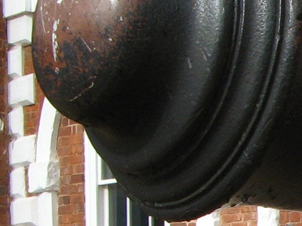 Close-up photo of a weathered black metal bell with a textured surface, possibly taken to demonstrate the macro photography capabilities of the Canon PowerShot A710 IS camera. The bell is against a blurred background of a brick building with white windows.
