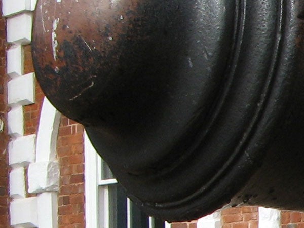 Close-up of a weathered outdoor bronze bell with a blurred red-brick building in the background.