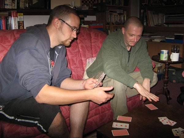 Two men sitting on a red sofa playing cards, with one holding a mobile device and both concentrating on the card game, in a living room with books and mugs in the background.