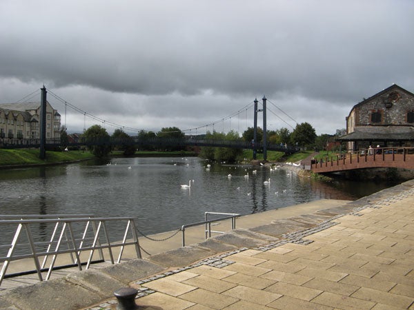 Photograph taken with a Canon PowerShot A710 IS showing a cloudy day at a serene riverside with swans, a pedestrian path, a small bridge, and old buildings in the background.