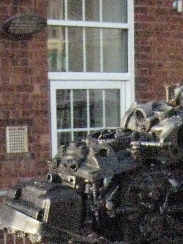 The image features a blurry shot of a brick building with a white-framed window, with the foreground focus unclear and possibly on a mechanical object or debris. The image quality suggests issues with camera focus or motion blur, characteristics that might be discussed in a review of the Canon Digital IXUS 850 IS's performance in varying conditions.