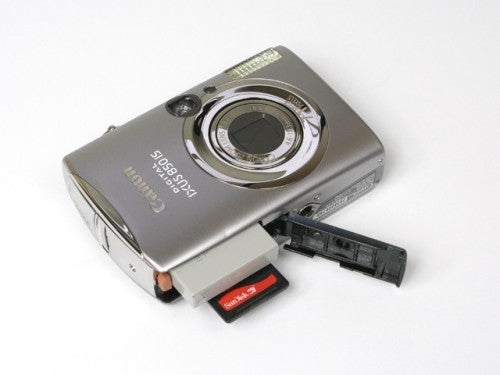 Canon Digital IXUS 850 IS camera with battery compartment open and memory card inserted, lying on a white surface.