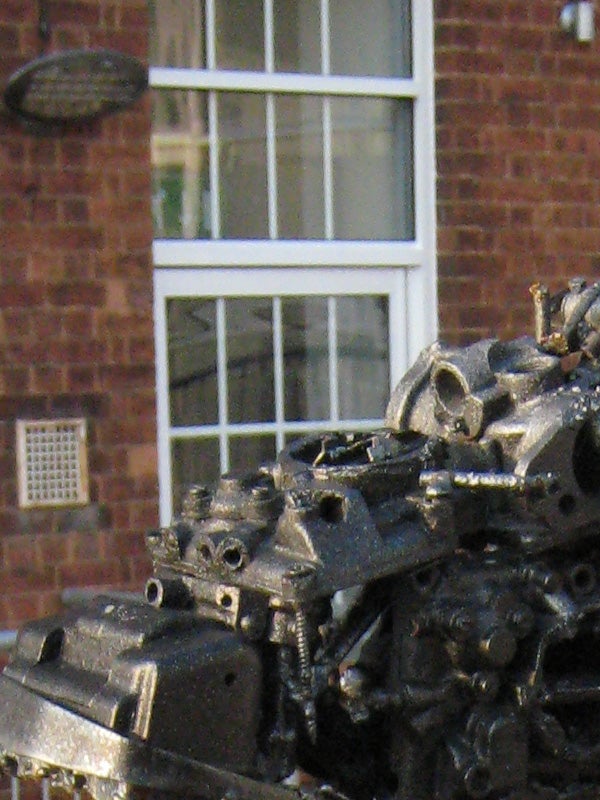 A close-up photograph displaying the detailed texture of a mechanical object, with a blurred building facade and windows in the background, demonstrating the shallow depth of field capability of the Canon Digital IXUS 850 IS camera.