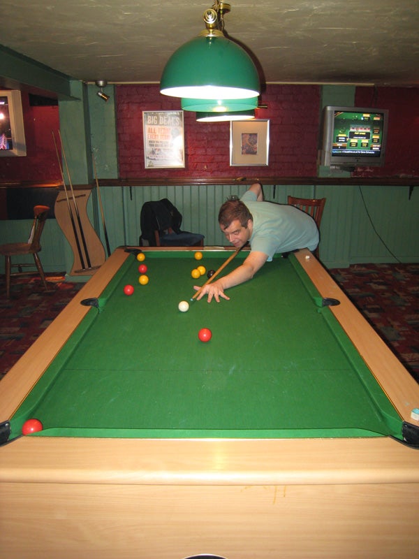A person plays a game of pool under a green hanging lamp in a room with red walls and posters.