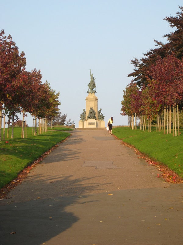 A clear photograph depicting a person walking on a pathway towards a statue monument framed by trees with autumn colored leaves against a blue sky, likely taken with a Canon Digital IXUS 850 IS camera.