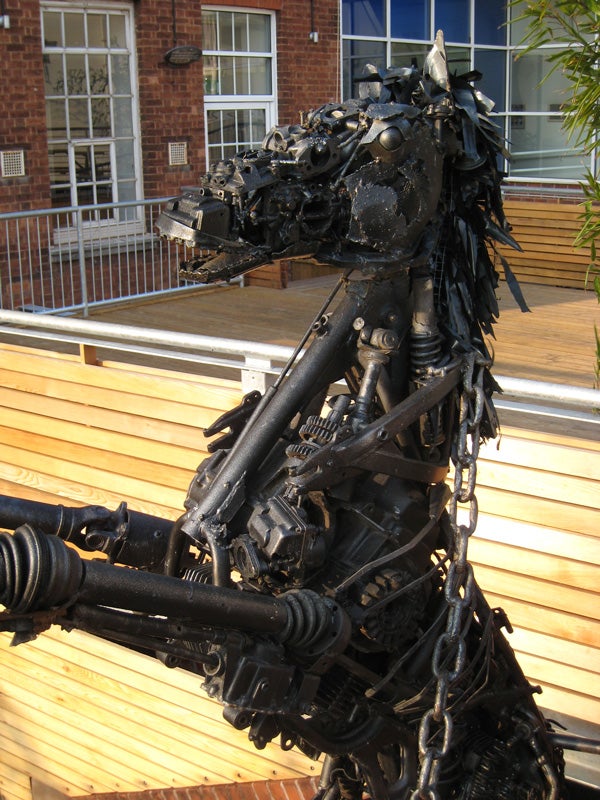 Sculpture of a horse made from assorted metal parts including gears and chains, displayed outdoors on a wooden bench with a brick building in the background.
