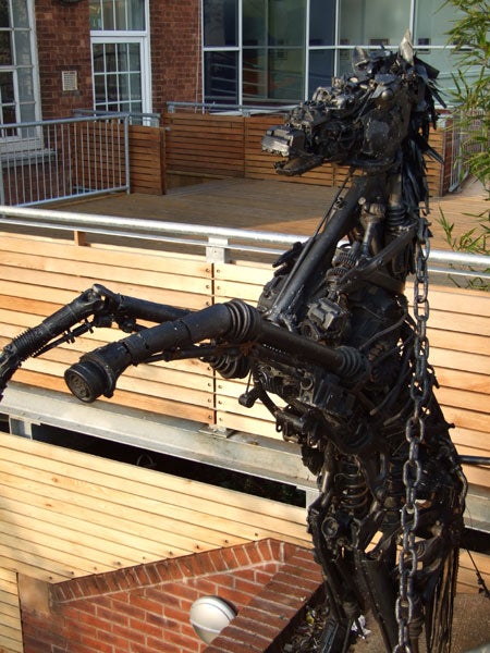 Metal sculpture of a dinosaur made out of mechanical parts displayed outdoors on a wooden bench