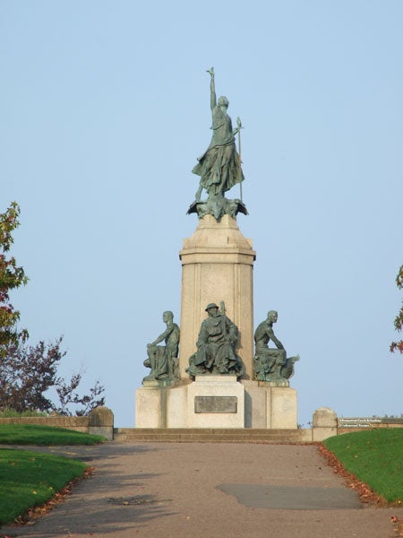 The image depicts a war memorial statue with multiple figures on a pedestal against a clear sky, situated at the end of a paved pathway with grass and trees to the sides.