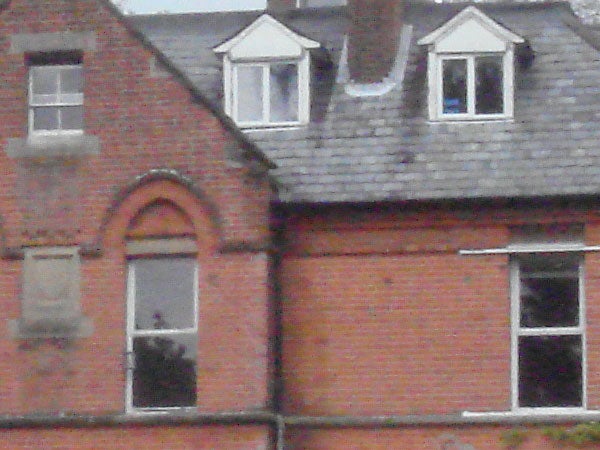 The image shows a close-up of a brick building with various windows. There are dormer windows on the roof and an arched window on the second story, indicating a traditional architectural style. The focus of the image is not sharp, potentially illustrating the image quality when using a digital camera like the Sony Cyber-shot DSC-T10 under specific conditions.
