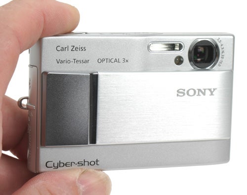 A hand holding a Sony Cyber-shot DSC-T10 digital camera, showcasing the front view with the lens retracted and the camera turned off. The camera features a Carl Zeiss Vario-Tessar lens with a 3x optical zoom, displayed prominently on the top right of the camera's silver body.