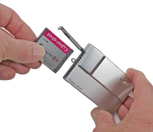 A person inserting a Memory Stick Pro Duo card into the slot of a silver Sony Cyber-shot DSC-T10 digital camera.