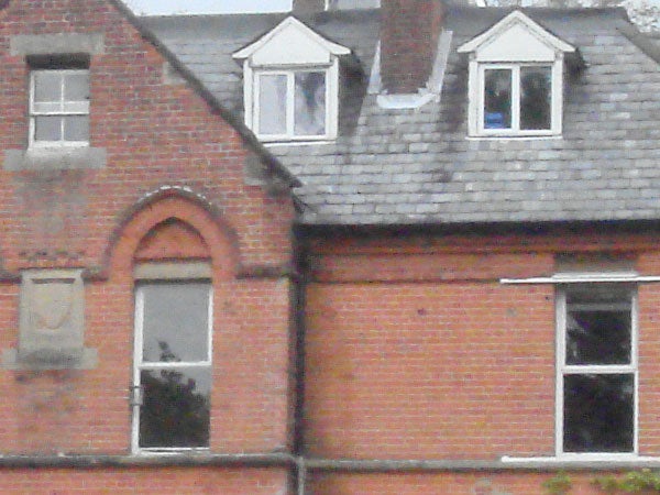 The image shows a brick building with multiple windows and roof dormers. The photo appears grainy and lacks sharpness, indicative of a high digital zoom level or a lower resolution sensor.
