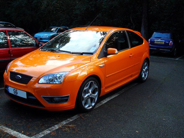 Orange Ford Focus parked on the side of a street with trees and other parked cars in the background.