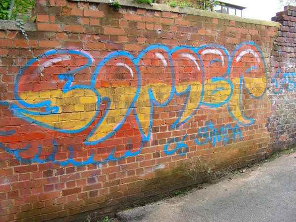 The image depicts a colorful graffiti artwork on a brick wall. The graffiti features bold lettering in blue and yellow with a signature or initial 