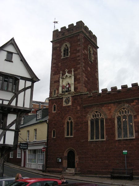 Old brick church tower with a clock and a small group of figures at the top, possibly taken with a Nikon Coolpix S10 camera. Traditional half-timbered house and modern buildings in the background under an overcast sky.