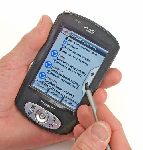 Hand holding a Mio DigiWalker P550, displaying navigation instructions on the screen with a stylus pointing to the display.