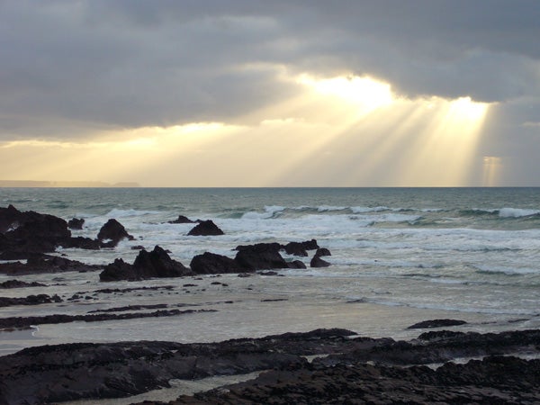 A serene seascape with sunlight streaming through clouds onto a rocky shoreline with tumultuous waves, showcasing the image quality of the Panasonic Lumix DMC-FX3 camera.