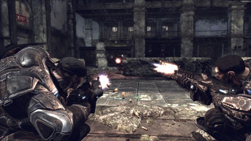 A screenshot from the video game Gears of War showing two characters in combat, taking cover behind a wall and firing their weapons at an off-screen enemy. The environment is a gritty, war-torn building interior.