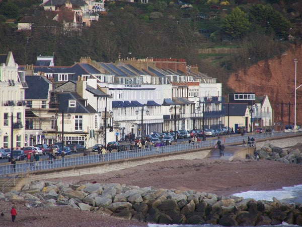 Photograph taken with Kodak EasyShare Z710 showing a clear view of a seaside promenade with people and cars, a pebble beach in the foreground, and a line of houses and buildings backed by green hills.