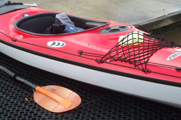 A red and white kayak with black netting on the deck next to an orange paddle, possibly demonstrating the Kodak EasyShare V705 Dual Lens Camera's ability to capture vibrant colors and fine details in outdoor lighting conditions.