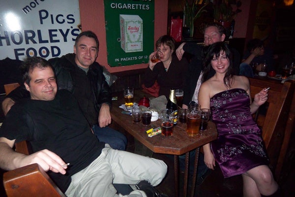 Group of people enjoying drinks at a pub with friends, with pints of beer on the table, ashtrays, and a 'no smoking' sign in the background; likely taken with a wide-angle setting demonstrating the capabilities of a camera like the Kodak EasyShare V705 Dual Lens.