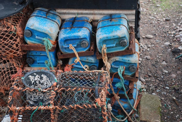 Blue weathered petrol containers stacked beside a lobster trap with orange netting, both resting on a pebbled surface.