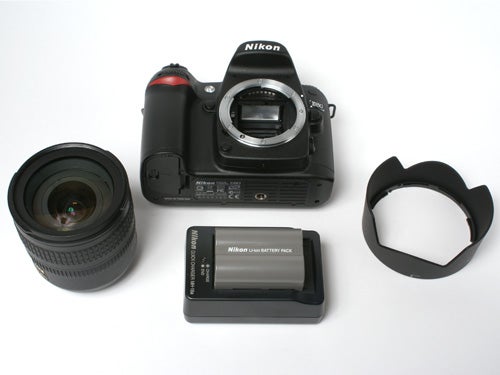 Nikon D80 DSLR camera body without a lens attached, accompanied by a detached lens, a lens hood, and a battery pack, all arranged on a white surface.