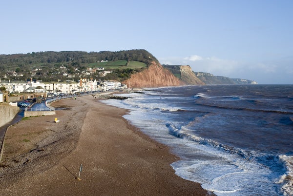 A scenic landscape photograph showing a beach view with waves hitting the shore, a small town to the left, and a prominent cliff in the background under a clear blue sky.