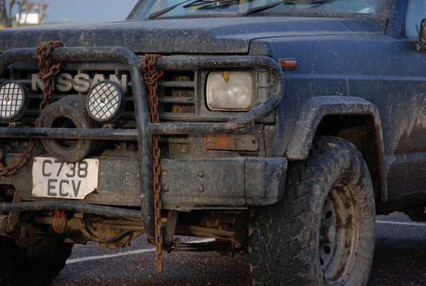 Close-up of a dirty and mud-splattered Nissan truck front end, showcasing the vehicle's grille, headlight, and license plate.