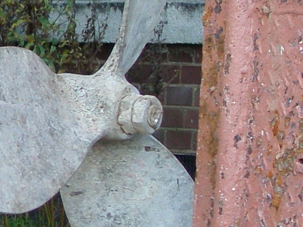Close-up image of a large, weathered propeller blade against a part-brick and part-plaster wall, showcasing the detail captured by an Olympus mju 1000 10MP Compact Camera.