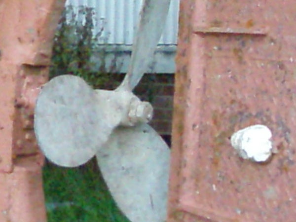 Close-up of a weathered and rusted propeller mounted on brickwork with paint chipping away, displaying signs of deterioration and age.