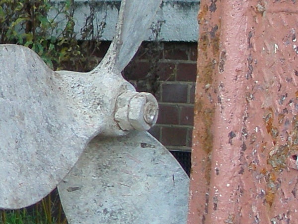 Close-up of an aged boat propeller next to a weathered brick wall.