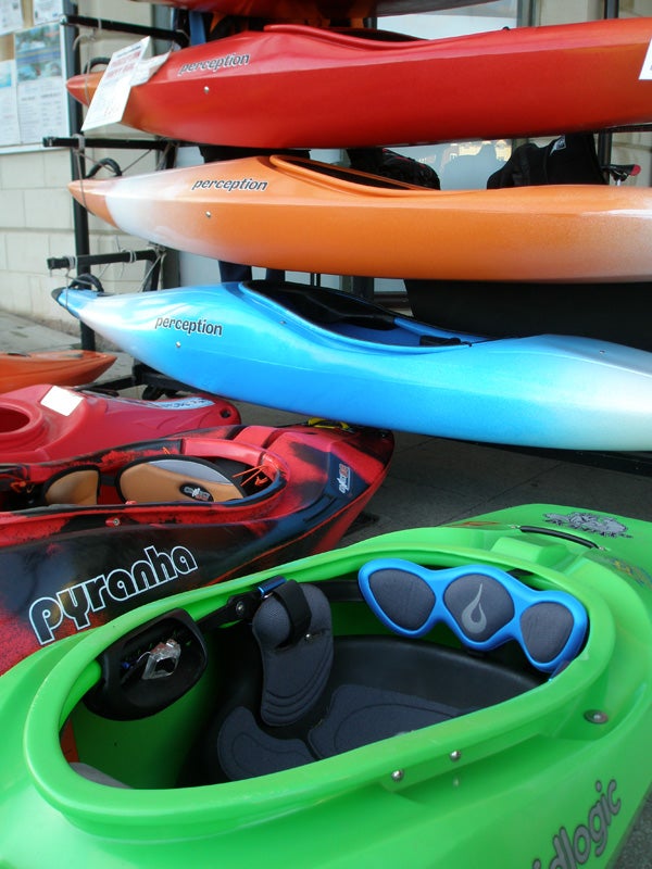 A selection of colorful kayaks stacked on a display rack, including brands like Perception and Pyranha, with a focus on a vibrant green kayak in the foreground.