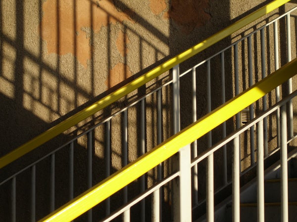 A sharp and detailed photograph demonstrating the image quality of the Olympus mju 1000 10MP Compact Camera, featuring the contrasting shadows and textures of a yellow railing against a concrete wall.