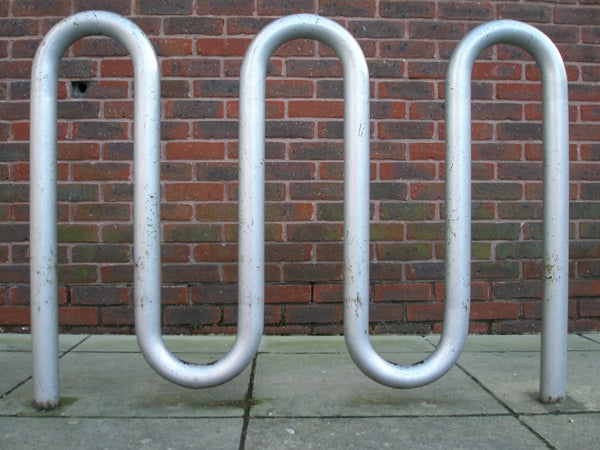 Three metal bike racks in front of a brick wall on a paved surface.