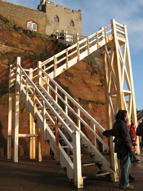 A wooden staircase leading up to a historic building on top of an orange cliff, with several people standing and looking at the view, bathed in warm sunlight.