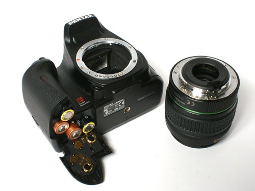 A Pentax K100D digital SLR camera body with an open battery compartment next to a detached camera lens on a white background.