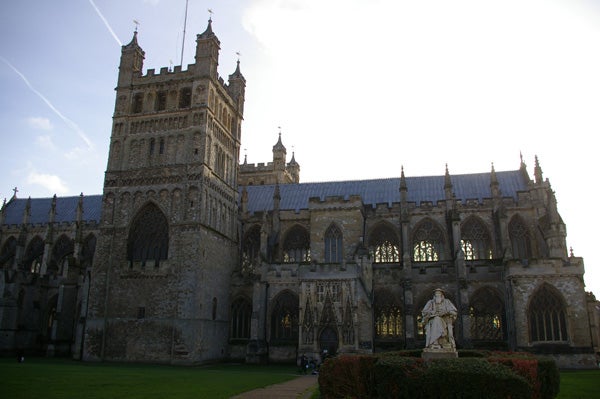 Photograph of an historic cathedral exterior taken in the sunlight showing intricate architectural details and a statue in the foreground on a clear day.