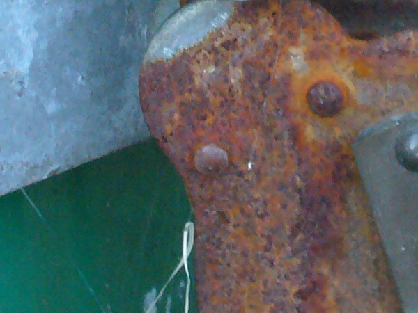 The image displays a close-up of a heavily rusted metal surface with two bolt heads and part of a green object.