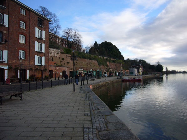 A serene riverside promenade with a row of old brick buildings on the left, a clear sky overhead, and calm waters reflecting the surroundings, likely captured with a Panasonic Lumix DMC-FX01 camera.