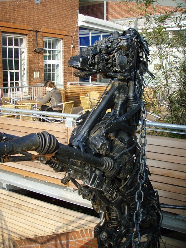 Sculpture of a horse constructed from mechanical parts and scrap metal displayed on a wooden deck with a person seated in the background.
