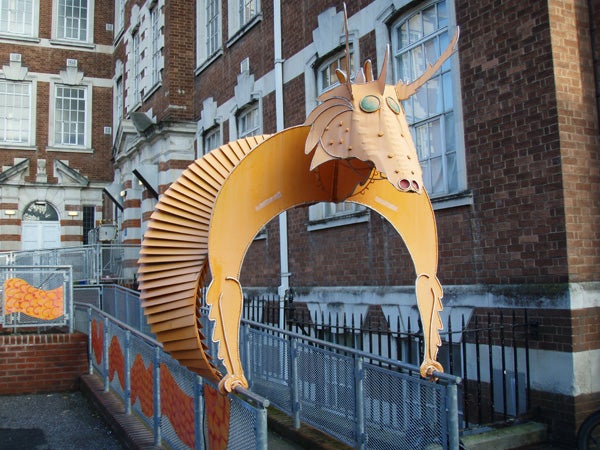 The image shows a large orange mechanical sculpture of a goat with articulated segments and green circular elements representing eyes, situated in an urban environment against a backdrop of a traditional brick building.