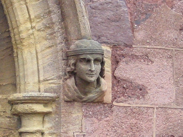 Stone sculpture of a human face with a headband carved into the exterior wall of a sandstone building.