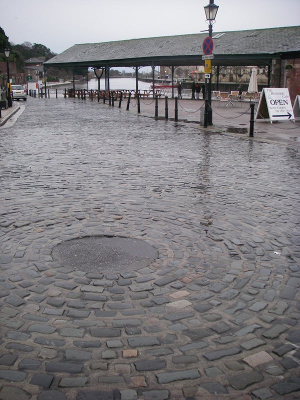 Wet cobblestone street with a puddle reflecting the surroundings, captured in cloudy weather, exemplifying the water-resistant capabilities of the Pentax W20 camera.
