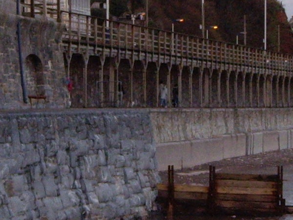 Image taken with a Pentax Optio T20 camera showing an overcast scene of a seaside promenade with a stone balustrade and pedestrians in the distance. There's visible graininess and loss of detail likely due to low light conditions.