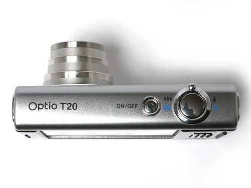 Pentax Optio T20 digital camera positioned horizontally on a white surface, featuring a silver body with a retractable lens, the model name clearly printed on the front, and a power button along with other control elements visible on the top panel.
